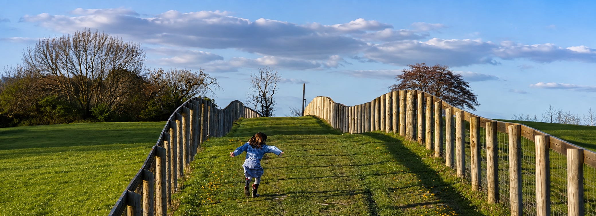 A child running up a fenced grass path through fields near Henley-on-Thames in golden evening light