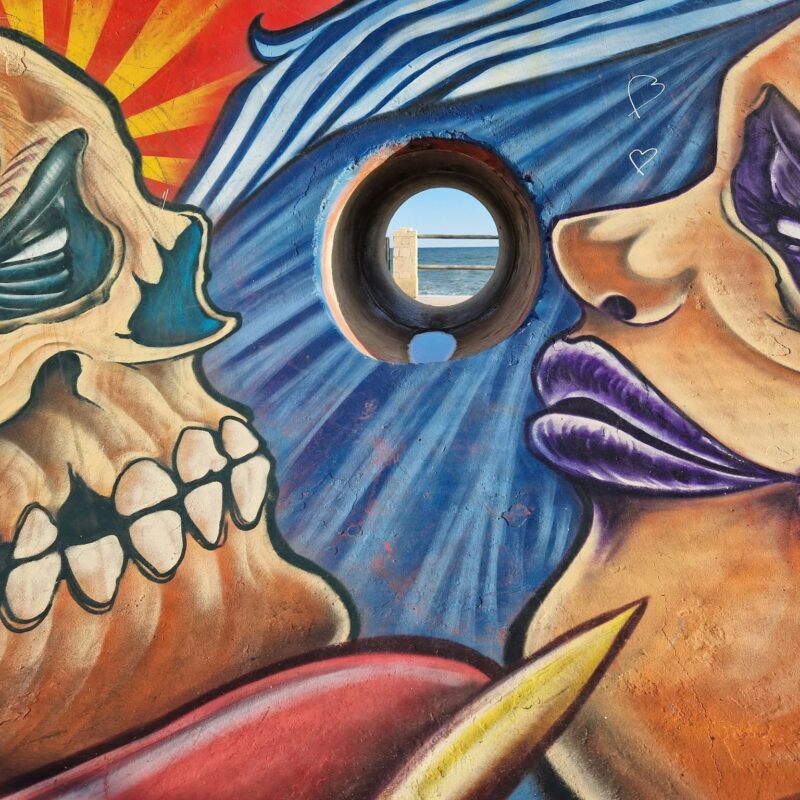 Street-art mural of two skull faces turned towards each other, with a round porthole framing the sea between them