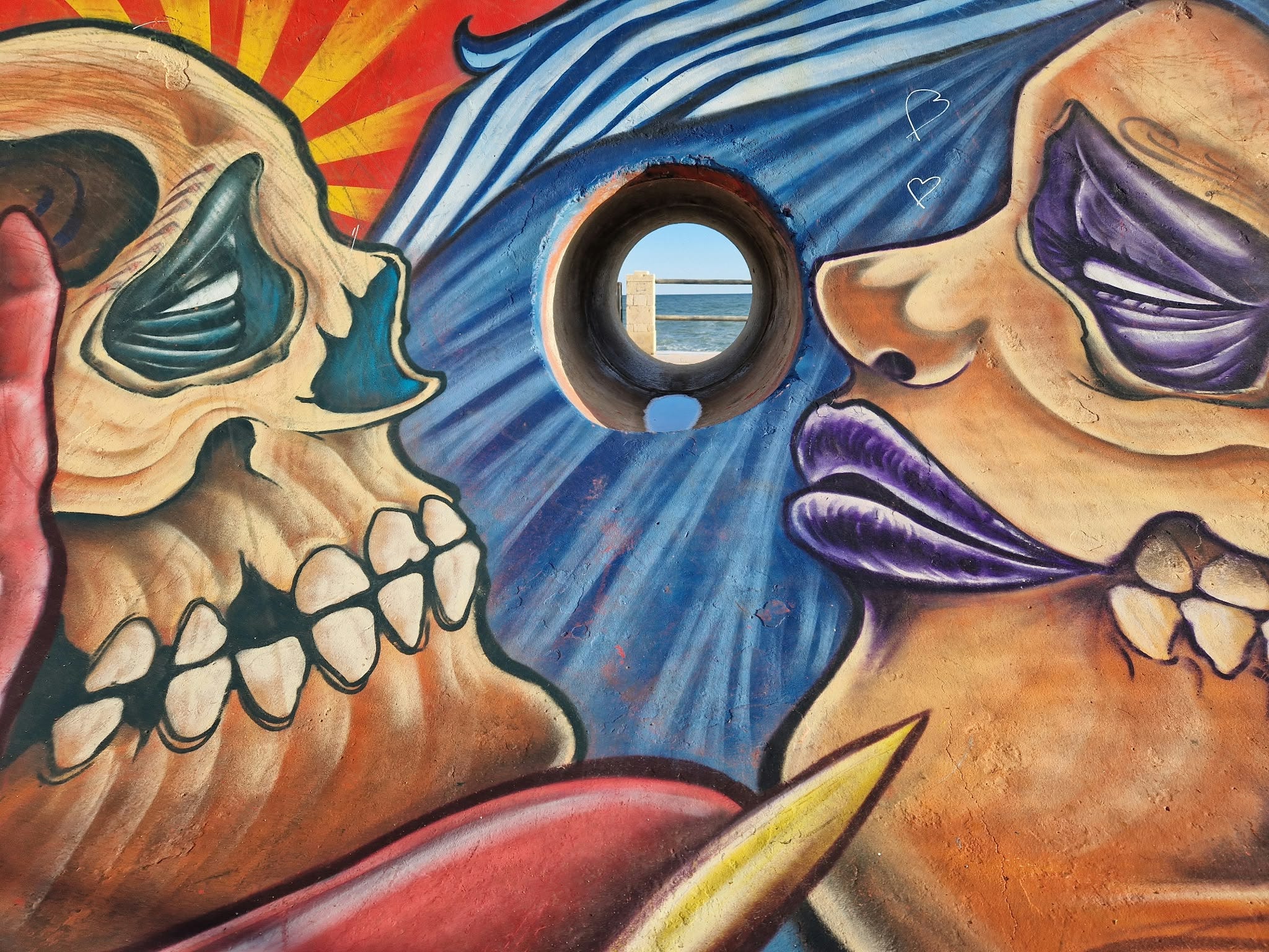 Street-art mural of two skull faces turned towards each other, with a round porthole framing the sea between them