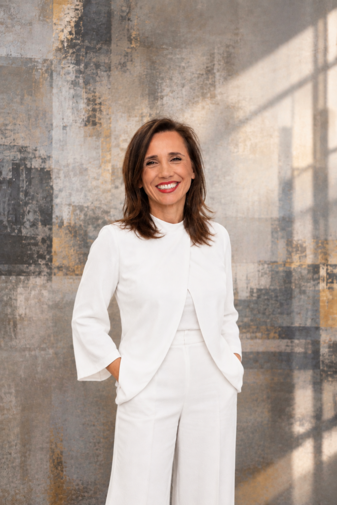 Livia Bernardini, smiling, in a white jacket and trousers, standing against a textured grey wall